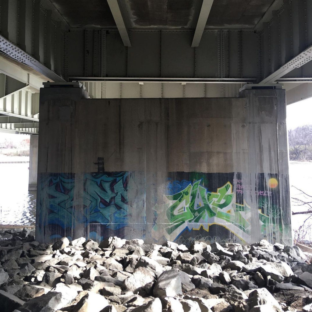 Faded graffiti under a bridge. Rocks in the foreground, water in the background.